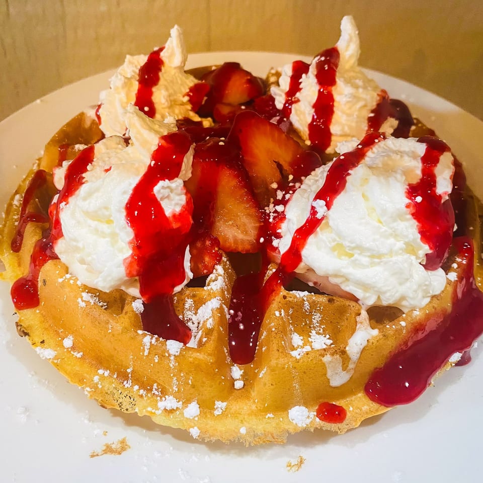 Golden waffle topped with whipped cream, strawberry slices, red syrup, and powdered sugar.