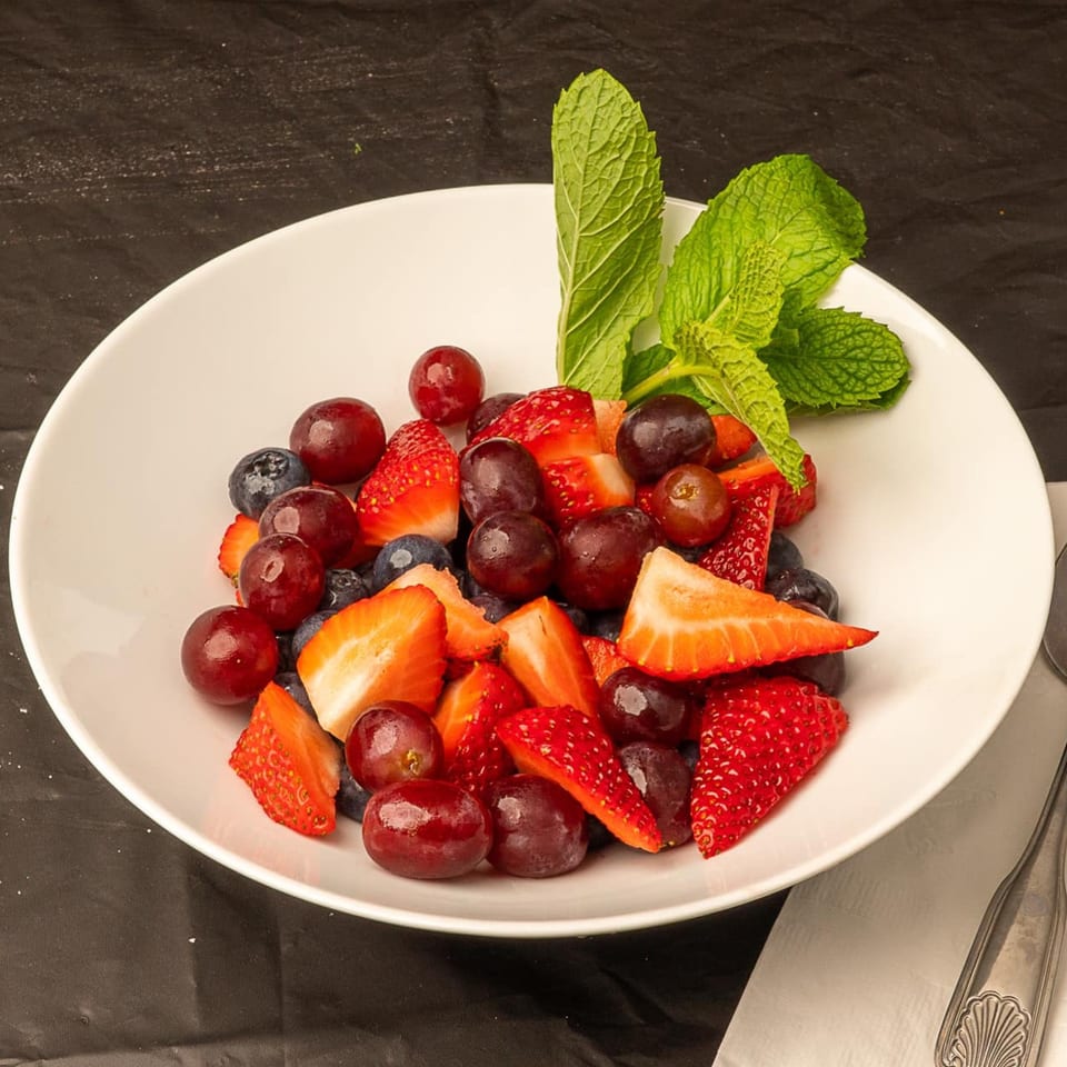 A white bowl filled with fresh fruit, including strawberries, blueberries, and red grapes, garnished with mint.